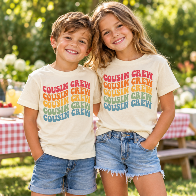 Two children wearing natural Cousin Crew Wavy shirts with five colorful rows of retro lettering at a family picnic.