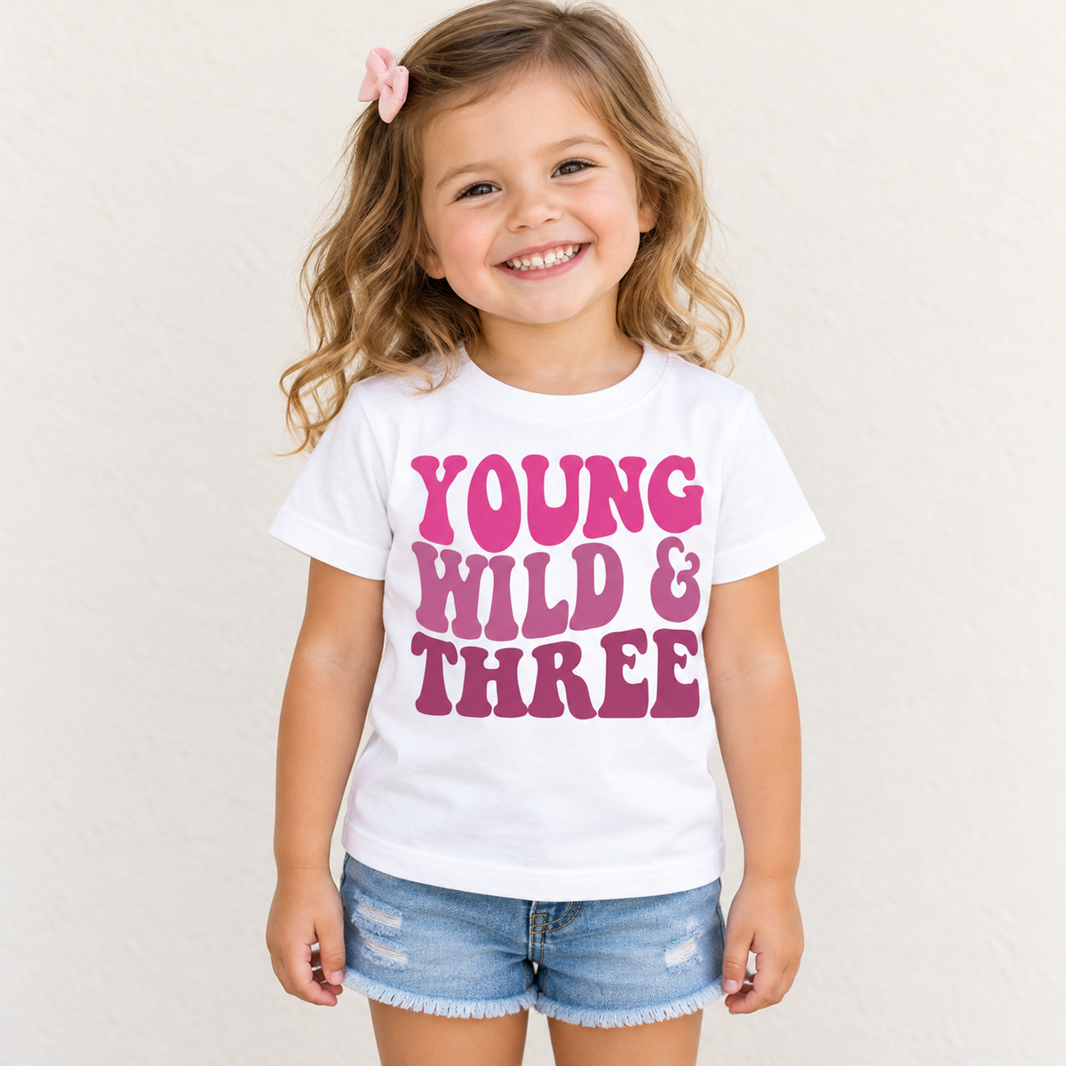 Girl wearing a white Young Wild and Three birthday shirt with pink retro lettering against a simple cream background.