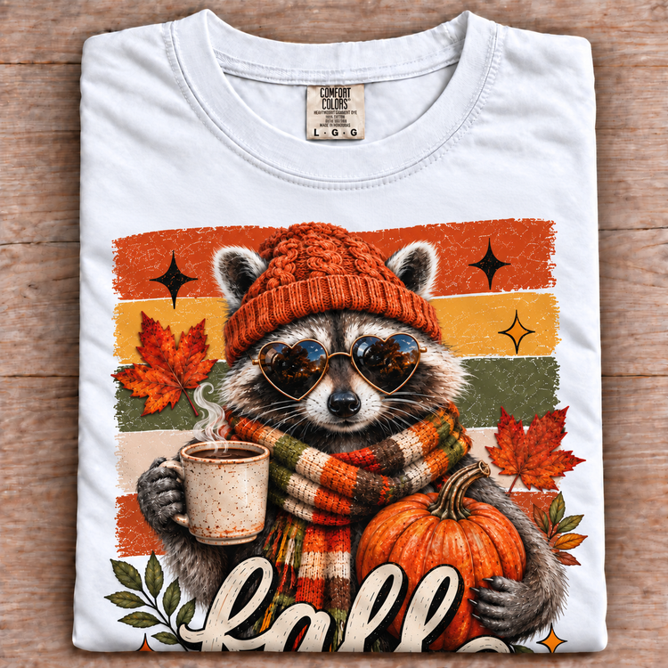 White Comfort Colors fall raccoon shirt with pumpkin and coffee graphic folded on wood