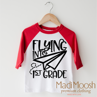 Flying Into First Grade Back To School design on a red-accented shirt.