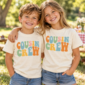 Two children wearing natural Cousin Crew shirts with colorful retro lettering at an outdoor family picnic.