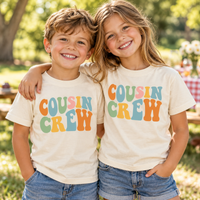 Two children wearing natural Cousin Crew shirts with colorful retro lettering at an outdoor family picnic.