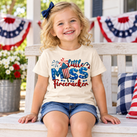 Toddler girl wearing a natural Little Miss Firecracker tee on a porch decorated for July Fourth.