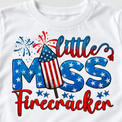 Close-up of Little Miss Firecracker lettering, stars and striped rocket artwork on a white cotton tee.