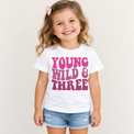 Girl wearing a white Young Wild and Three birthday shirt with pink retro lettering against a simple cream background.