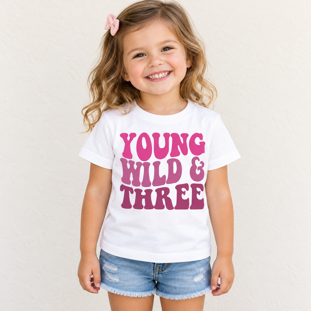 Girl wearing a white Young Wild and Three birthday shirt with pink retro lettering against a simple cream background.